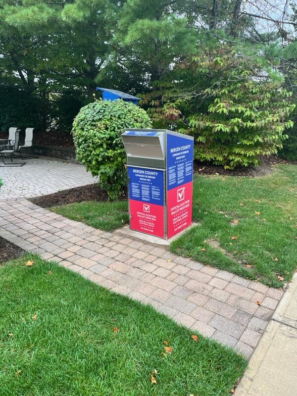 A photo of the newly installed ballot box. It is along a brick pathway and is red on the bottom and blue on the top with white text.