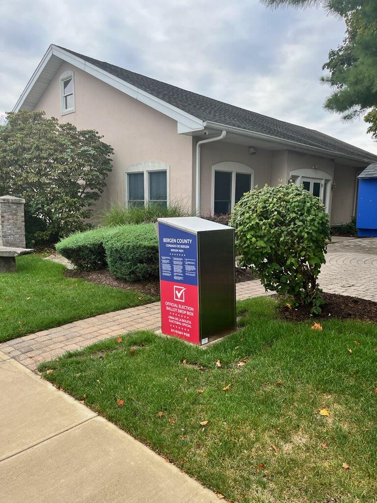 A photo of the newly installed ballot box from the back side of it. It is along a brick pathway and is red on the bottom and blue on the top with white text.